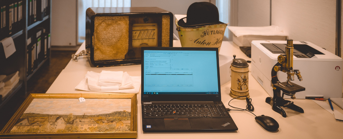 Laptop mit geoeffneter Datenbank zur Erfassung von Objekten. Um den Laptop herum stehen mehrere Museumsobjekte. Ein Gemälde, ein altes Radio, ein Mikroskop.