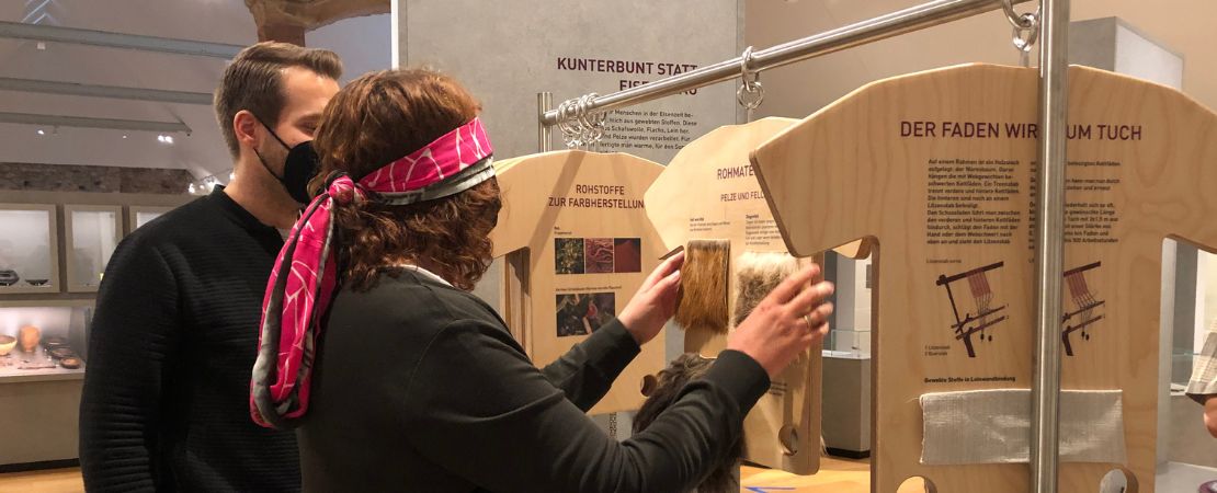Eine Frau mit verbundenen Augen und ein Mann stehen in einer Ausstellung vor einer Kleiderstange. An der Kleiderstange haengen Holztafeln in T-Shirt-Form. Die Frau beruehrt textile Materialien auf der Holztafel.
