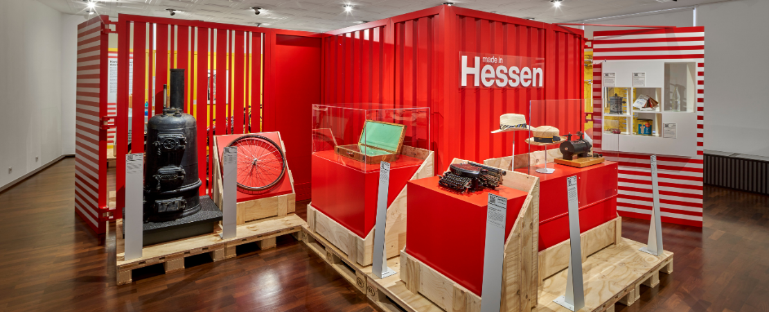 Ausstellungsansicht der Ausstellung Made in Hessen. Vor einer roten Container-Wand stehen mehrere Exponate auf Paletten wie Fahrradreifen, Schreibmaschine.