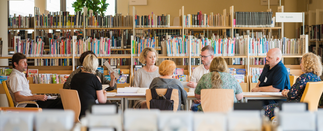 Eine Gruppe von Menschen sitzt verteilt an einem langen Tisch in einer Bibliothek. Im Hintergrund stehen volle Buecherregale.