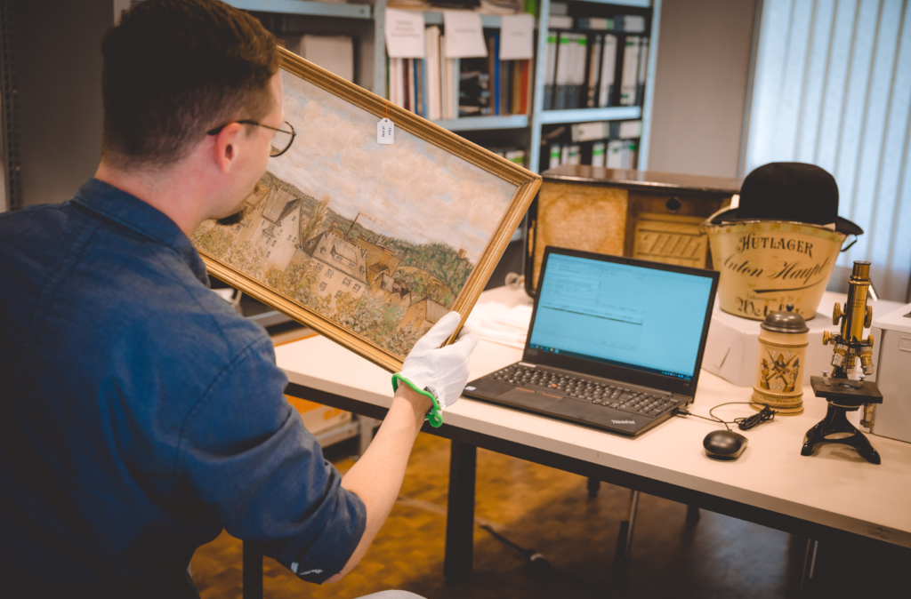 Mann hält Gemälde in der Hand. Auf einem Tisch steht ein geöffneter Laptop mit Datenbank. Dahinter weitere Museumsobjekte.