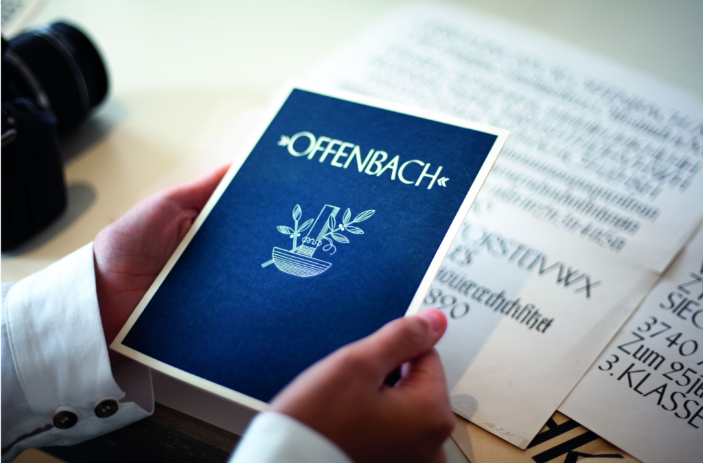 Eine blaue Karte mit weißem Schriftzug "Offenbach" wird von zwei Haenden gehalten.