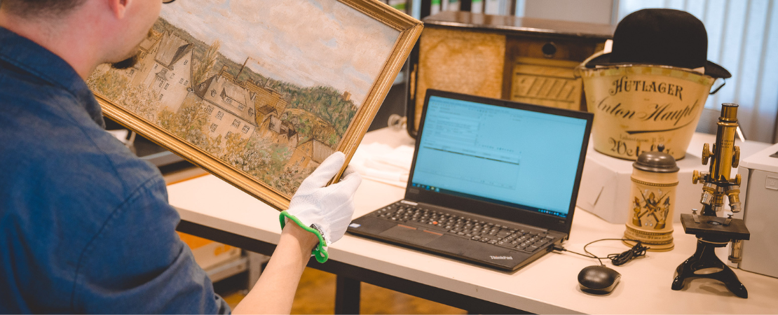 Mann hält Gemälde in der Hand. Auf einem Tisch steht ein geöffneter Laptop mit Datenbank. Dahinter weitere Museumsobjekte.
