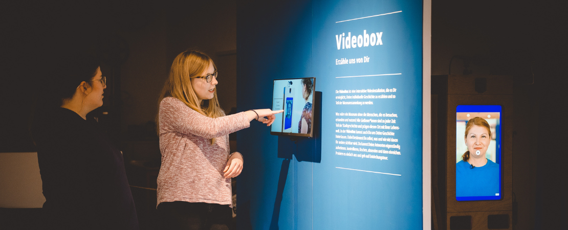 Zwei Personen stehen vor einer Video-Installation. Die Person rechts zeigt auf den Bildschirm. Im Hintergrund ein Screen mit Person auf Bildschirm.
