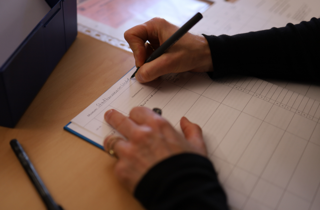 In der rechten Hand haelt eine Person einen Stift und schreibt "Stadtmuseum Wetzl..." in ein Eingangsbuch. Die linke Hand liegt neben auf.