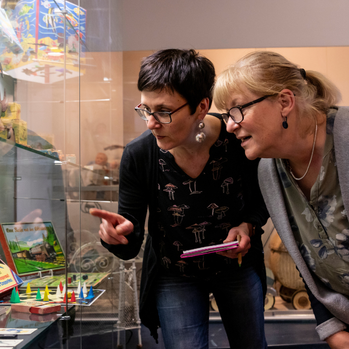 Zwei Frauen stehen gebeugt vor einer Glasvitrine und schauen gespannt auf ein Objekt, eine zeigt in die Richtung.
