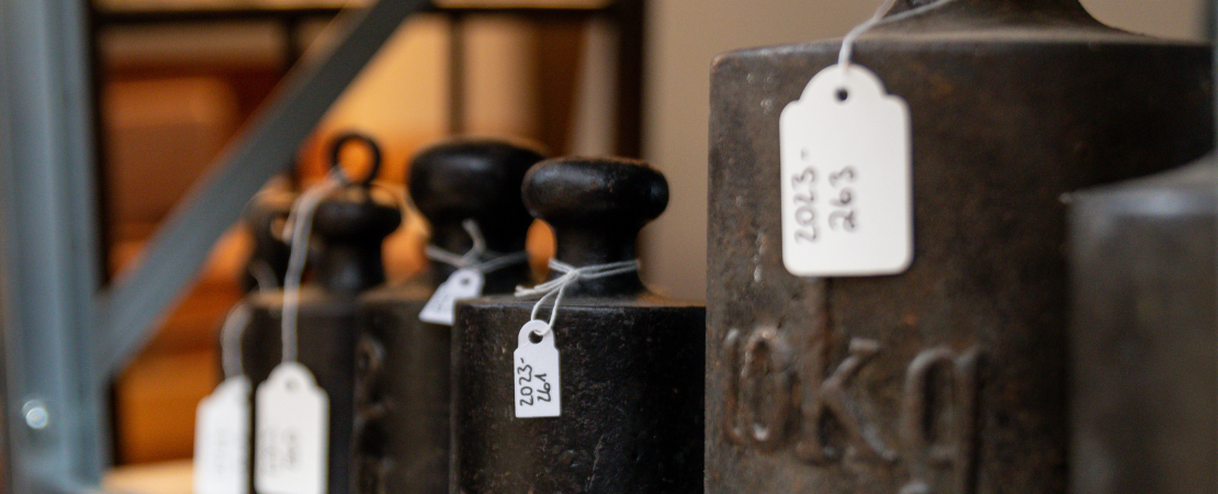 Gewichte in unterschiedlichen Größen stehen aufgereiht in einem Regal und sind jeweils mit einem Schild mit zugehöriger Inventarnummer gekennzeichnet.