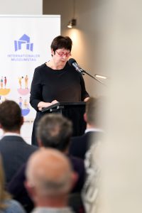Dr. Birgit Kümmel am Rednerpult bei ihren Grußworten anlässlich der Veranstaltung. Im Hintergrund ein Banner des Internationalen Museumstags.