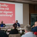 Vortragssituation: Die Pop-Up-Ausstellung, die den Verbandstag 2024 begleitet, wird dem Publikum und dem Vorstand vorgestellt. Menschen sitzen vor einer Leinwand. Auf der Leinwand ist ein magentafarbenes Quadrat zu sehen, auf dem steht: Quality-Time mit der eigenen Sammlung?