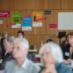 Blick auf die Pop-Up-Ausstellung, die den Verbandstag 2024 begleitete. Im Vordergrund sitzen Die Teilnehmenden des Verbandstags. Die Wand im Hintergrund zeigt den Schriftzug "Date im Depot. Alte Schätze neu entdeckt". Daneben sind beispielhafte Plakate angebracht, die Beispiele zur Objektinszenierung präsentieren.