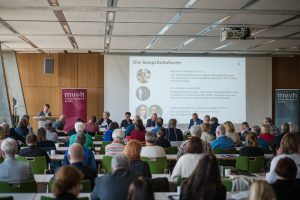 Vortragssituation. Im Vordergrund ist das Publikum mit den Hinterköpfen zu sehen, im Hintergrund an der Leinwand befindet sich eine Projektion mit dem Titel "Die Gesprächsforen". Darunter werden kurz die Leiterinnen der drei Gesprächsforen mit Poträtfoto vorgestellt. Vor der Leinwand sitzen zwei weitere Personen, dem Publikum zugewandt. Links im Bild steht eine Person am Rednerpult und spricht.