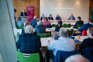 Ansicht mit Publikum beim Grußwort von Staatsminister Timon Gremmels. Am Rednerpult steht der Staatsminister, daneben sitzt dem Publikum der geschäftsführende Vorstand und Christina Reinsch. Im Hintergrund ist eine Präsentationsfolie mit dem Titel "Grußwort Staatsminister Timon Gremmels" zu erkennen.