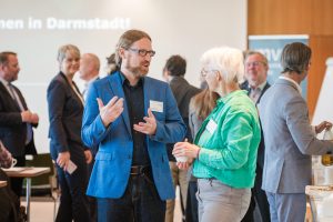 Eine Gruppe von Menschen steht in einem Konferenzraum. Im Vordergrund unterhalten sich ein Mann mit Brille, Bart und blauem Sakko und eine Frau mit weißen Haaren und grünem Hemd angeregt miteinander. Im Hintergrund stehen weitere Teilnehmer in kleinen Gruppen und unterhalten sich ebenfalls.