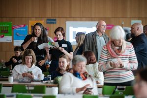 In einem Konferenzraum stehen mehrere Teilnehmer in Gruppen beisammen. Im Vordergrund sitzt eine Frau in einem gestreiften Pullover und betrachtet ihr Smartphone, während andere Menschen im Hintergrund in Gespräche vertieft sind und Broschüren lesen.