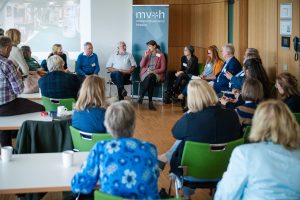 Circa 20 Personen sitzen auf Stühlen im Kreis und sind ins Gespräch vertieft. Im Hintergrund steht ein RollUp, auf dem das Logo des Museumsverbands Hessen abgebildet ist.