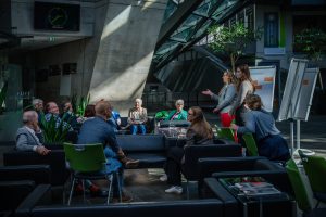 Im Foyer des Darmstadtiums, eines großen Veranstaltunsgebäudes in Darmstadt, fand das Gesprächsforum von Laura Cabanus und Luisa Wipplinger statt. Beide stehen vor einer Gruppe sieben Personen, die in Sesseln sitzen und unterhalten sich über das Thema Schaudepot. Hinter den Gesprächsleiterinnen befinden sich drei Flipcharts, an denen das Gespräch dokumentiert wird.