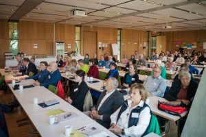 Ansicht der rund hundert Teilnehmenden, die an ca. sechs Tischreihen sitzen und in Richtung Rednerpult blicken.