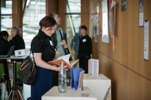 Blick auf die Pop-Up-Ausstellung, die den Verbandstag 2024 begleitete. Eine Teilnehmerin Schaut sich die Beispiele an und blättert in einer ausgestellten Publikation. An einer Wand sind Karten in Form eines Smartphones angebracht, die Beispiele für Objektinszenierung bei Social Media präsentieren. Vor der Wand befindet sich eine Stele, auf der ein Tablet weitere Beispiele präsentiert werden.