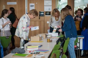 Im Vordergrund ist eine Frau zu sehen, die sich von einem Tisch mit ausgelegten Materialien einen Flyer nimmt und darin liest. Um sie rum sind weitere Personen zu erkennen, die sich teilweise unterhalten.