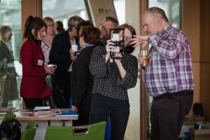 Zwei Personen betrachten eine Smartphone-Halterung zur Foto-Stabilisierung. Eine Person hält die Halterung in der Hand und zeigt auf etwas auf dem Smartphone. Im Hintergrund sind weitere Personen zu erkennen.
