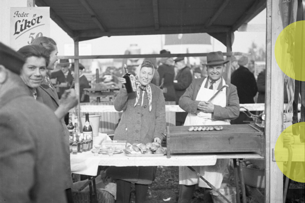 Eine historische Schwarz-Weiß-Aufnahme eines Marktstandes. Eine Frau in einem Mantel und mit Kopftuch hält eine Flasche in die Kamera. Neben ihr steht ein Mann mit Schürze und Hut, der mit einer Zange vor einem Grill mit Würstchen steht. Im Hintergrund sind weitere Marktbesucher zu erkennen. Ein Schild mit Aufschrift wird halb von einer Person im Vordergrund verdeckt.
