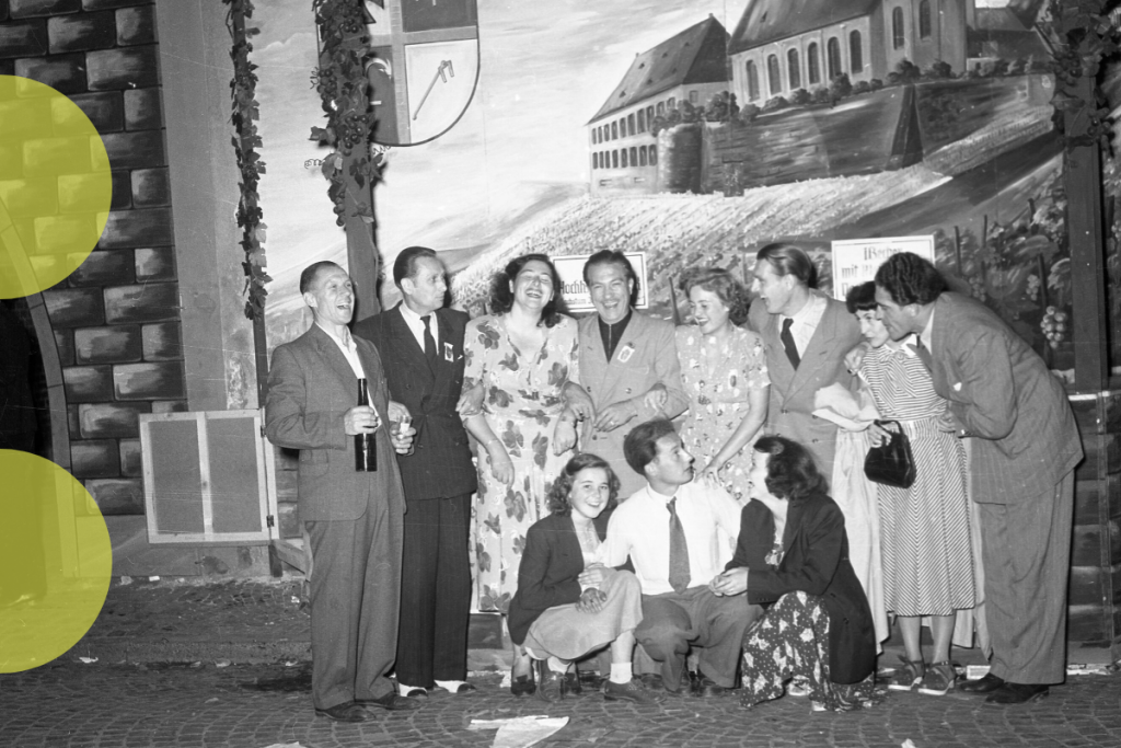 Ein Schwarz-Weiß-Foto einer Gruppe von Männern und Frauen, die in festlicher Stimmung sind. Einige der Personen lachen, andere halten Flaschen oder Gläser in den Händen, während zwei Frauen und ein Mann in der vorderen Reihe auf dem Boden sitzen. Im Hintergrund ist ein gemaltes Wandbild mit einer Weinlandschaft und einem Wappen zu sehen.