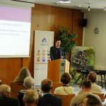 Dr. Daniel Groth, Concultura steht hinter einem Stehpult. Dahinter ist das Banner zum Internationalen Museumstag sowie eine Leinwand zu sehen. Im Vordergrund sitzen Gäste der Veranstaltung in Stuhlreihen mit Sicht auf den Redner.