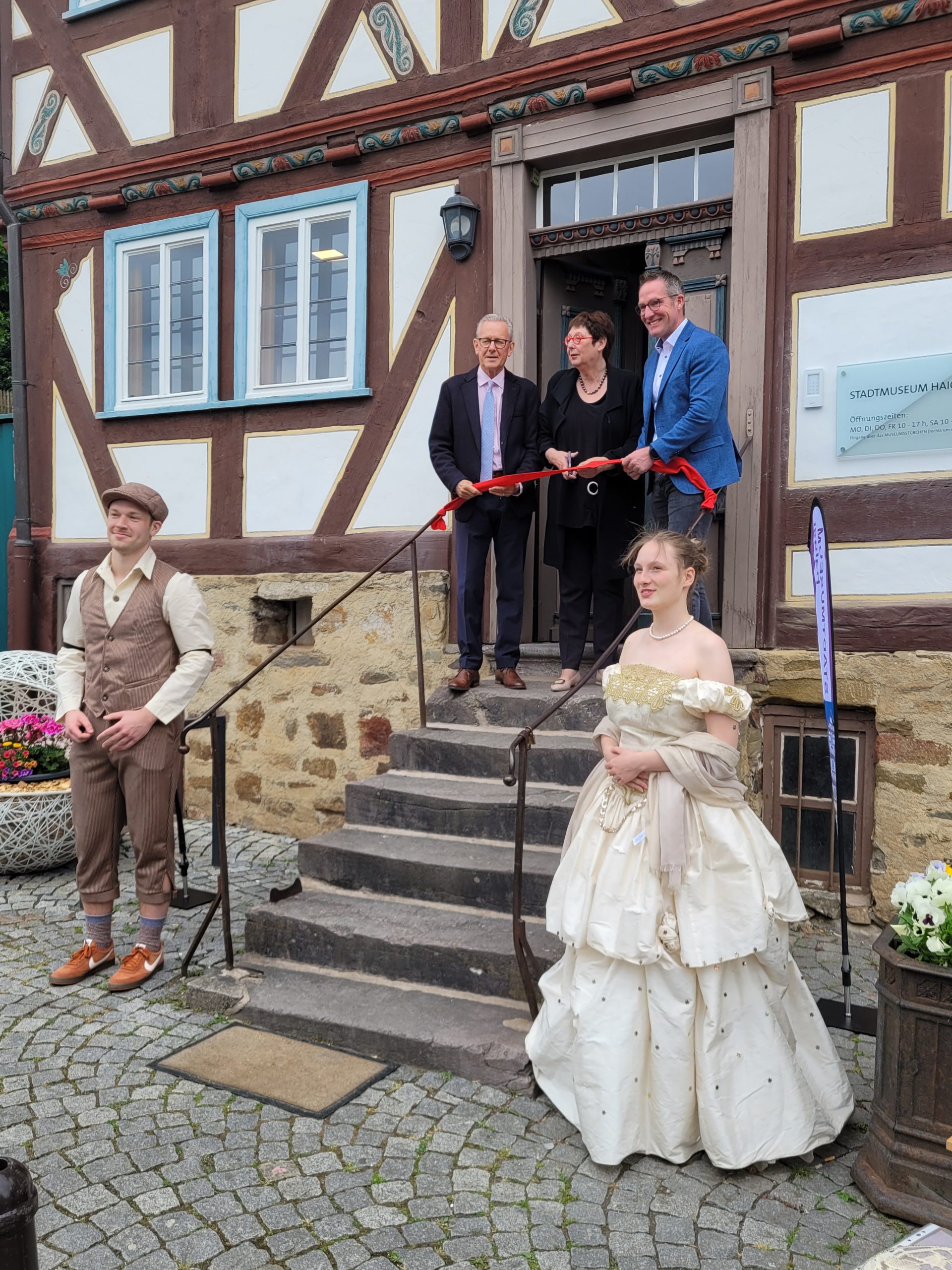 Eine Treppe führt zum Eingang des Museums. Davor stehen in historischer Kleidung die "Maskottchen" Isabella und Eduard. Vor dem Eingang schneiden v.l.n.r Bürgermeister Mario Schramm, Vorsitzende Dr. Birgit Kümmel und Landrat Carsten Braun ein rotes Band durch.