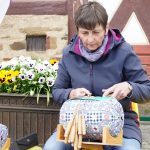 Eine Frau sitzt vor dem Museumsgebäude auf dem Stuhl und klöppelt.