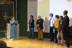 sieben Personen stehen auf einer Bühne, links Person mit Mikro, im Hintergrund Leinwand