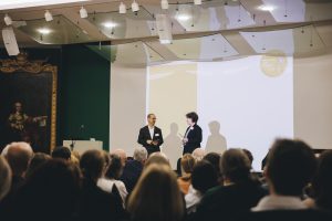 Zwei Personen stehen auf einer Bühne, davor Zuschauende von hinten fotografiert, im Hintergrund Leinwand mit Schattenwurf der Personen.