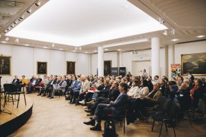Blick in einen großen Saal der gut gefüllt ist, etwa acht Stuhlreihen, alle schauen nach vorne, im Hintergrund Gemälde, vorne links eine kleine Bühne mit Stühlen erkennbar.