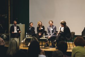 sechs Personen sitzen auf einer Bühne, vorne Tische mit Getränken, eine Person mit Mikro in der Hand stellt gerade eine Frage, die anderen schauen aufmerksam.