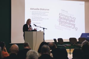 Person steht an einem Redner*innenpult mit Mikro, im Hintergrund eine PowerPoint eingeblendet. im Vordergrund Zuschauende.