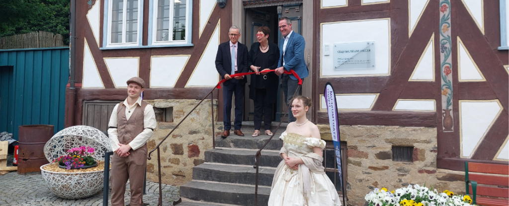 Drei Menschen stehen auf der Treppe vor dem Eingang zum Stadtmuseum und schneiden ein rotes Band durch Am unteren Ende der Treppe stehen zwei Personen in historischer Kleidung
