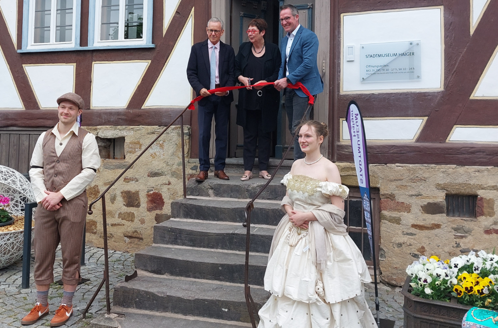 Eine Treppe führt zum Eingang des Museums. Davor stehen in historischer Kleidung die "Maskottchen" Isabella und Eduard. Vor dem Eingang schneiden v.l.n.r Bürgermeister Mario Schramm, Vorsitzende Dr. Birgit Kümmel und Landrat Carsten Braun ein rotes Band durch.