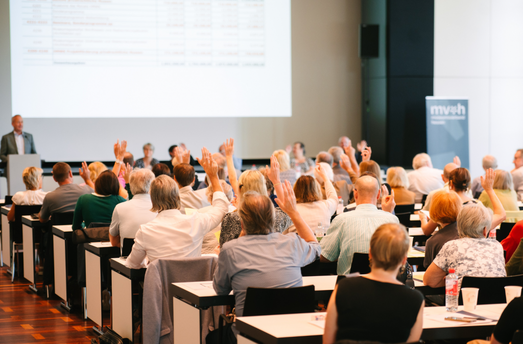 Menschen sitzen in einem Konferenzraum und heben die Hand, während vorne eine Person am Rednerpult spricht. Es handelt sich um eine Versammlung oder Abstimmung mit zahlreichen Teilnehmenden.