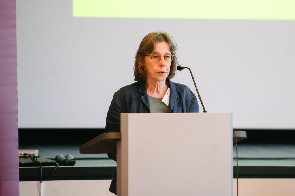 Dr. Bettina von Andrian Hessen steht an einem Rednerpult mit Mikrofon und spricht Richtung Publikum. Im Hintergrund ist ein dunkelroter Banner des Museumsverbandes mit Logo und eine Leinwand zu sehen.