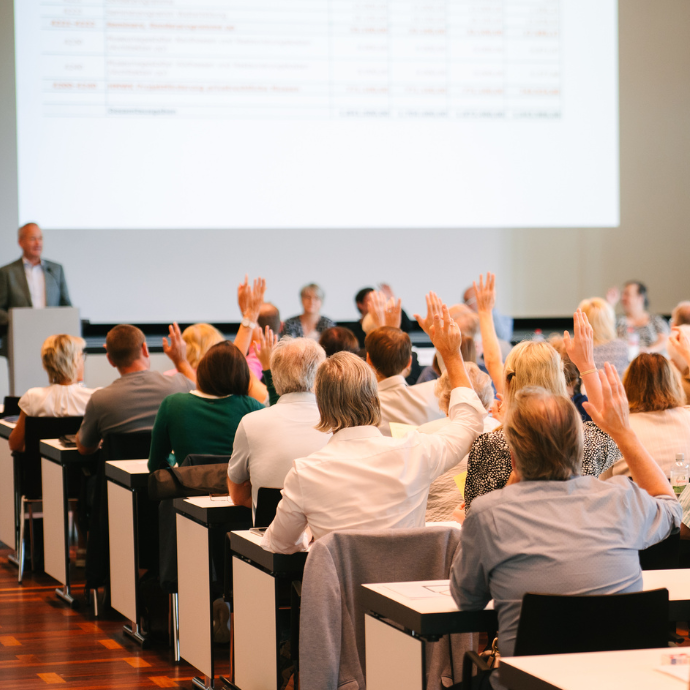 Menschen sitzen in einem Konferenzraum und heben die Hand, während vorne eine Person am Rednerpult spricht. Es handelt sich um eine Versammlung oder Abstimmung mit zahlreichen Teilnehmenden.