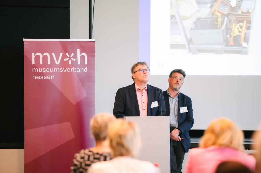 Frank Gnegel und Dr. Joel Fischer stehen an einem Rednerpult mit Mikrofon und sprechen Richtung Publikum. Im Hintergrund ist ein dunkelroter Banner des Museumsverbandes mit Logo und eine Leinwand zu sehen.