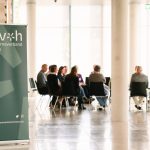 Gruppe von Personen sitzt in einem hellen Raum im Stuhlkreis und nimmt an einer Gesprächsrunde teil. Im Vordergrund ist ein grüner Banner des Museumsverbandes Hessen zu sehen.