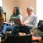 Eine Gruppe von Personen in einem Stuhlkreis. Ein Mann arbeitet an seinem geöffneten Laptop, eine Frau hat ein Mobilgerät in der Hand. zwei andere Frauen blicken in Richtung der Gesprächsleitungen.