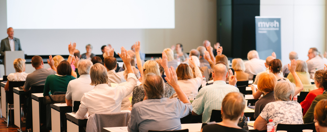Menschen sitzen in einem Konferenzraum und heben die Hand, während vorne eine Person am Rednerpult spricht. Es handelt sich um eine Versammlung oder Abstimmung mit zahlreichen Teilnehmenden.