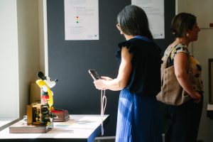 Eine Person fotografiert mit ihrem Smartphone eine Micky-Mouse Figur, die ein Telefon in der Hand hält, auf einem Tisch stehend.