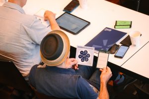 Ein Mann mit Hut hält zwei Karten in der Hand, vor ihm auf einem Tisch liegen eine Tagungsmappe, ein Portemonnaie, ein Trinkbecher und ein Smartphone.