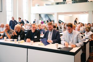 Fünf männliche Personen sitzen in einer Tischreihe nebeneinander und schauen sich teilweise an. Darunter unter anderem Staatsminister Timon Gremmels, der eine Tagungsmappe des Museumsverbandes Hessen öffnet. Im Hintergrund sind viele Teilnehmende in Tischreihen zu sehen.