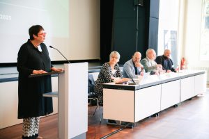 Dr. Birgit Kümmel steht sprechend am Rednerpult mit Mikrofon. Neben dem Rednerpult sitzen fünf Personen in einer Tischreihe bestehend aus Vorstandspersonen und Christina Reinsch am Beginn der Tischreihe.