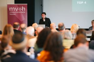 Dr. Birgit Kümmel am Rednerpult den Teilnehmenden zugewandt. Im Vordergrund sind verschwommen viele Personen von hinten mit Köpfen zu erkennen. Im Hintergrund ist eine Leinwand mit Projektion und Personen zu sehen. Links neben dem Rednerpult steht ein dunkelroter Banner des Museumsverbandes Hessen mit Logo.