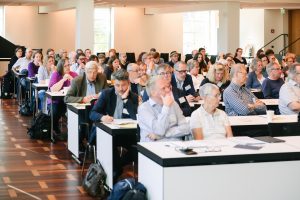 Menschen sitzen in einem Konferenzraum in Tischreihen und schauen Richtung Bühne bzw. Rednerpult.