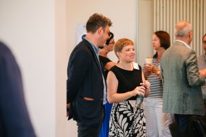 Eine Gruppe von Personen die sich unterhält. Im Vordergrund sind ein Mann und eine Frau, die sich unterhalten und lächeln.